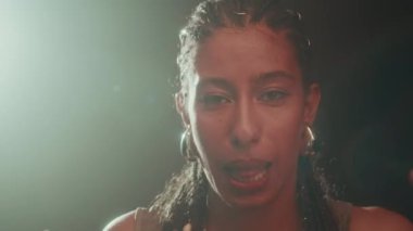 Saçları örgülü, metal küpeleri olan, koyu arkaplanda ışık kaynağıyla kameraya hip hop dansı yapan, kendinden emin, çift ırklı bir kız.