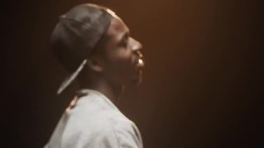 Kendine güvenen, kasketli, kolyeli siyah bir adam gece partisinde yanıp sönen ışıkta dans ediyor.