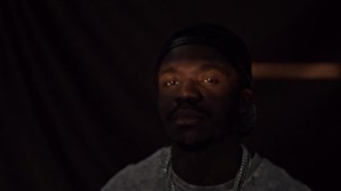 Beyzbol şapkası ve zincirli kolye takan özgüvenli Afro-Amerikalı adamın göğüs portresi. Gözlerinde ve yüzünde kısmi ışık demeti var. Görüntülerin geri kalanı karanlıkta.
