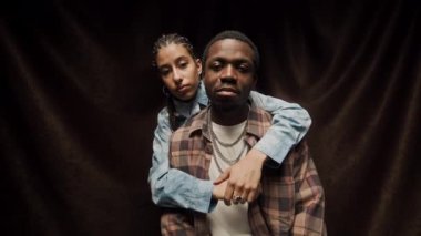 Siyah arka planda kameraya poz veren Afro-Amerikan havalı genç çiftin orta boy portresi. Saçları örgülü bir kız, erkek arkadaşını arkadan kucaklıyor.