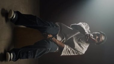 Siyah şapkalı, kolyeli, büyük beden tişörtlü ve bol kotlu Afrikalı bir adamın karanlık odada projektör ışığının altında oturup rap yaptığı dikey bir fotoğraf.