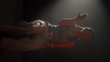 Kendine güvenen, zincirli kolye takan, sandalyede oturmuş rap yapan ve elleri koyu arkaplanda hip hop hareketleri yapan bir kızın dikey çekimi.