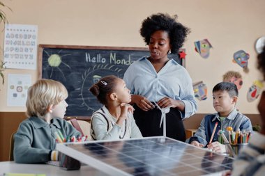 İlkokuldaki çoklu etnik kökenli çocuklar güneş paneliyle büyük bir masada oturup siyah etnik kökenli bayan öğretmenlerin sınıfta yenilenebilir enerji hakkında konuştuklarını dinliyorlar.