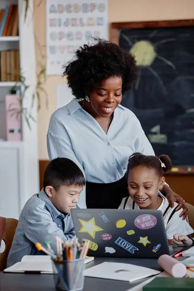 stock image Vertical shot of smiling female teacher of Black ethnicity talking to primary students looking at laptop screen