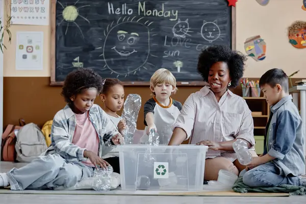 stock image Multiethnic group of children and female teacher in ecology club sorting waste putting plastic bottles in recycling bin in classroom