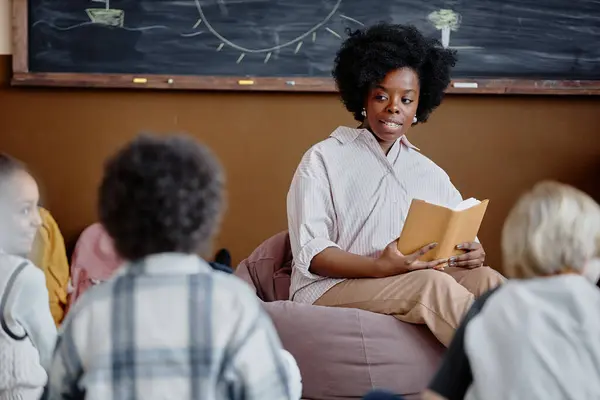 Young female teacher of Black ethnicity sitting in bean bag chair against blackboard reading captivating story aloud to kids around in classroom, copy space