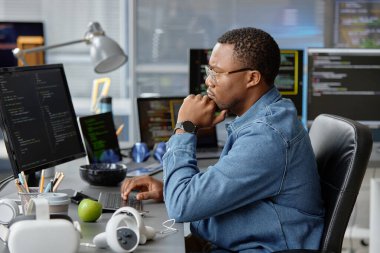 Çalışma masasında birden fazla bilgisayar ekranı olan, son teknoloji oyun geliştirme için kod yazan düşünceli Afrikalı Amerikalı VR programcısının yan görüntüsü.