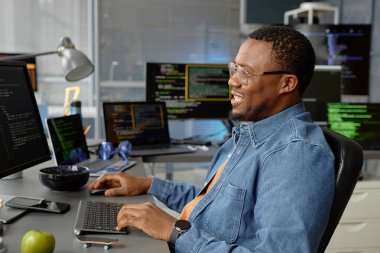 Siyah etnik kökenli gülümseyen erkek web geliştiricisinin yan görüntüsü. Çalışma masasında birden fazla ekranla otururken, ofiste kod programlama için bilgisayarı kullanarak, boşluğu kopyalayarak.