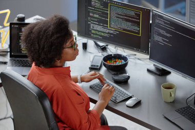 ,Masasında şeker kâsesi ile odaklanmış kıvırcık kadın Afrikalı Amerikalı yazılım geliştiricisinin ofis klavyesinde yazarken birden fazla bilgisayar ekranı incelemesi ve hata ayıklama kodunu okurken yüksek açılı görüntüsü.