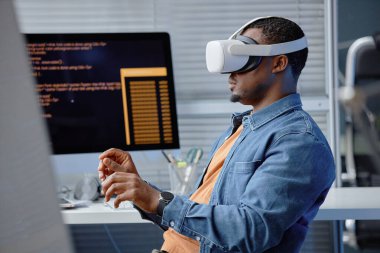 Erkek Afro-Amerikan yazılım mühendisinin VR uygulamasını test ederken ya da bilgisayar programcısı masasında otururken kulaklık takarken yan görüntüsü, boşluğu kopyala