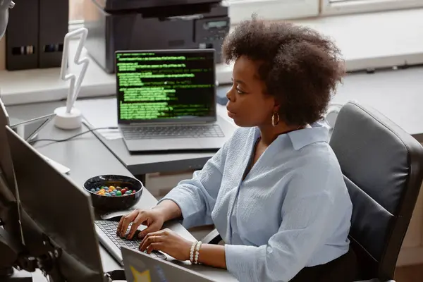 stock image High-angle side view shot of young focused African American software developer writing code on computer, while sitting at working desk with candy bowl at hand and multiple screens in modern office