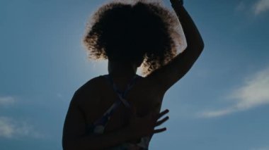 Kıvırcık saçlı, ellerini arkaya atıp, güneşli bir günde dışarıda antrenman yapmadan önce vücut hareketleri yapan, esneyen ve ısınan bir kadının silüetinin arka görüntüsü.