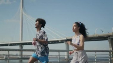 Aktif giyimli genç atletik çift, güneşli bir yaz gününde şehrin nehir kıyısında birlikte koşuyorlar. Arka planda ise şehir köprüsü.