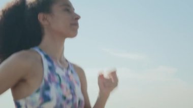 Spor sutyenli atletik kıvırcık saçlı bir kızın göğüs yukarı çekimi. Arka planda bulutsuz bir gökyüzü ile yaz günü açık havada koşuyor.