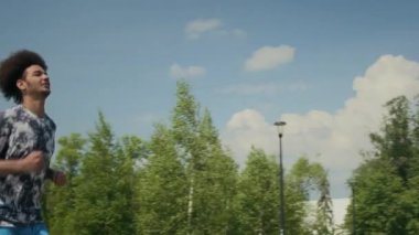 Spor kıyafetli genç bir adamın parkta koşarken, gülümserken ve kız arkadaşına beşlik çakarken yaz günü birlikte dışarıda egzersiz yaparken PAN yavaş çekim.