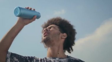 Genç kıvırcık saçlı adam yüzüne şişeden su dökerek ferahlatıyor ve dışarıda idmandan sonra dinlenirken kameraya bakıyor.