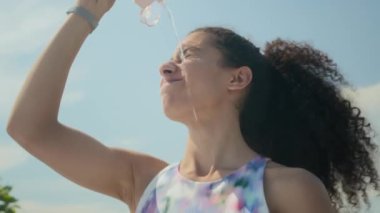 Genç sporcu kadın dışarıda duruyor, yüzüne su döküyor ve yoğun eğitimden sonra dinlenirken gülümsüyor.