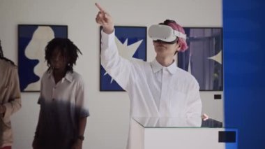 Pembe saçlı genç Asyalı adam modern sanat galerisinde interaktif turne sırasında VR kulaklığı kullanırken görünmez dokunmatik ekranda geziniyor.
