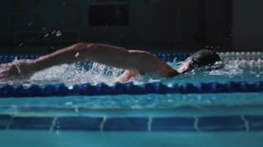Güçlü erkek yüzücünün yüzme şapkası ve gözlük takıp kapalı havuzda şeritlerle yarışırken çekilmiş yan görüntüsü.
