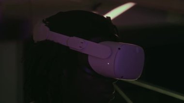 VR kulaklıklı genç bir adam karanlık neon ışıklı odada duruyor ve modern sanat galerisindeki ışık tesisatını ziyaret ederken kontrolörleri kullanıyor.