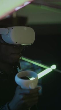 Siyah adamın VR gözlük ve kumanda kullanarak dikey çekimi. Karanlık salda neon ışığı kurulumu ve artırılmış gerçeklikteki kavramsal sanat galerisini keşfederken.