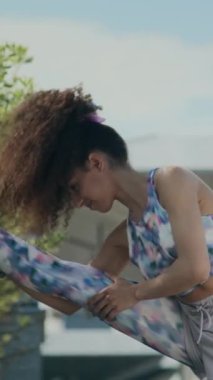 Spor giyimli kıvırcık saçlı bir kızın şehirde spor yaptıktan sonra bacak esnetme egzersizi yaptığı dikey çekim.