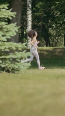 Genç bayan koşucunun kulaklıkla ve spor kıyafetleriyle parkta koşarken, yaz günü dışarıda egzersiz yaparken dikey çekimi.