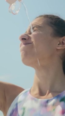 Genç, neşeli bir kadının yüzüne soğuk su dökerken ve sıcak yaz günlerinde açık hava eğitiminden sonra mutlu bir şekilde gülümserken dikey çekimi.