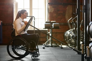 Spor salonunda tekerlekli sandalye kullanan ve halterle ağırlık çalışması yapan aktif bir kadının dramatik yan görüntüsü.