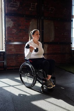 Spor salonunda tekerlekli sandalye kullanan ve günışığı ışınları tarafından aydınlatılan kuvvet eğitimi sırasında dambılı tutan yetişkin kadınların dramatik dikey portresi.