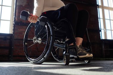 Spor salonunda tekerlekli sandalye kullanan ve güç eğitimi sırasında elinde dambıl tutan bir kadının güneş ışığı fotokopi alanının aydınlattığı motive edici yakın çekim.