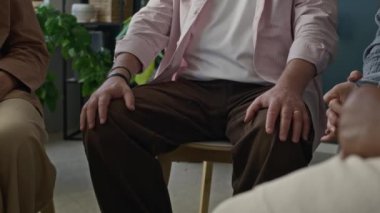 Cropped shot of unrecognizable man sitting with hands down on his knees and speaking during group therapy session with other people indoors