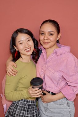 Vertical portrait shot of smiling young Hispanic girl embracing Asian female fellow student while looking at camera isolated on pink background clipart