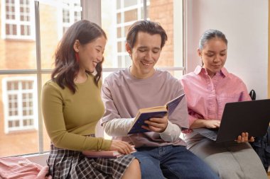 Genç adam ve Asyalı kız öğrenci birlikte gülüp kitap okurken İspanyol sınıf arkadaşları üniversitenin pencere kenarında dizüstü bilgisayarlarında sörf yapıyor.