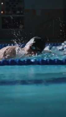 Dikey yan görüş, kapalı havuzda yüzerken serbest stil yüzme tekniği kullanarak kolunda dövme olan kararlı erkek sporcunun yavaş çekimini izliyor.