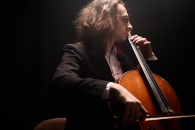 Orta ölçekli, uzun saçlı, çello çalan erkek müzisyenin karanlık stüdyoda ışık saçan klasik müzik konseri vermesi.