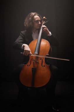 Düşey çekimde profesyonel erkek müzisyen tek başına çello çalıyor. Karanlık stüdyoda klasik müzik çalıyor.