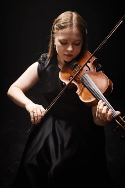 Profesyonel kadın kemancının dikey çekimi. Zarif siyah elbiseli. Karanlık stüdyoda üst ışık altında klasik müzik çalıyor.