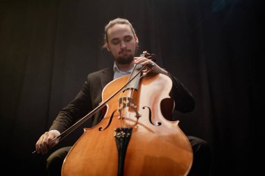 Projektör ışığı altında siyah perdelere karşı solo konserde çello çalan profesyonel müzisyenin düşük açılı çekimi.
