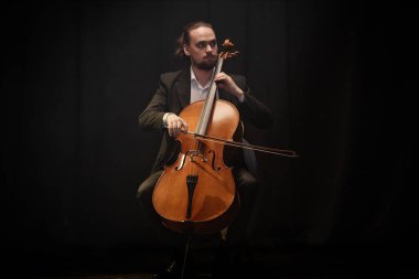 Solo konser sırasında çello çalan erkek klasik müzik sanatçısı projektör ışığı altında siyah perdelere karşı otururken, boşluğu kopyala