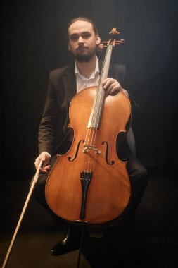 Dikey olarak, erkek klasik müzik çalar çello tutarken kameraya bakarken loş sisli stüdyoda projektör ışığı altında otururken.