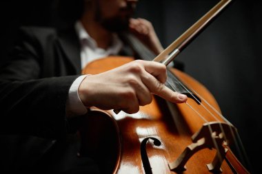 Siyah zemin üzerindeki kontrastlı ışıkta çello çalan tanınmamış erkek klasik müzik sanatçısının eline yaklaş, parmaklar ve tellere odaklan