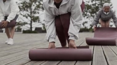 Yoga minderlerini parktaki ahşap zemine yuvarlayan üç yetişkin kadının yavaş çekimleri, güne açık havada yoga pratiğiyle başlıyorlar.