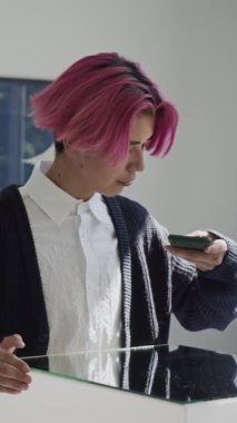 Dikey çekimde pembe saçlı Asyalı bir adam modern sanat galerisine giderken yansıtıcı ayna yüzeyinin akıllı telefonuyla fotoğraflarını çekiyor.