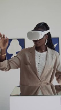 VR kulaklık takmış genç siyahi bir kadının modern sanat galerisinde görsel turlar yaparken havada görünmez nesnelere dokunuşunu dikey olarak büyüt.