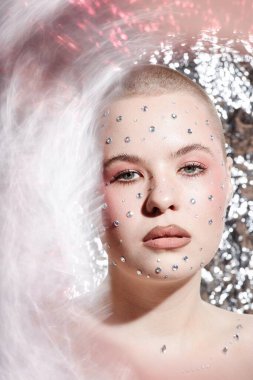 Vertical closeup portrait of young woman with creative makeup and gem stickers on face looking at camera on metallic background clipart