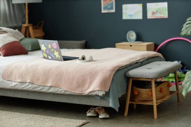 Rahat yataklı, yumuşak yastıklı modern yatak odası ve yatağın üzerine dizüstü bilgisayar. Rahat ve davetkar bir ortam yaratıyor. Duvarda doğal elementler ve çocuk çizimleri var.