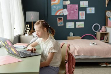 Down sendromlu genç bir kız yatak odasında oturmuş bilgisayarını online çalışmalar için kullanıyor ve etrafı hoş dekorlarla çevrili bir şekilde öğreniyor.