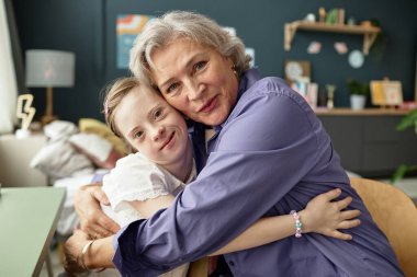 Keyifle Down sendromlu genç kıza sarılan yaşlı bir kadın. Sıcak bir odada. İkisi de sıcak bir şekilde gülümsüyor ve kişisel eşyalarıyla birlikte sandalyede yan yana oturuyor.