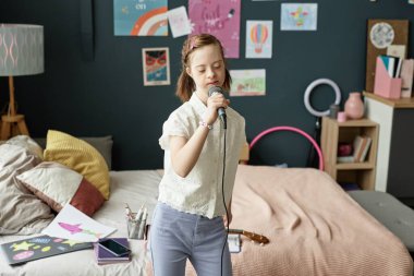 Yaratıcı dekorasyon ve çizimlerle çevrili, rahat yatak odasında mikrofona şarkı söyleyen genç bir kız. Pencereden gelen doğal ışık renkli ve davetkar boşlukları aydınlatır